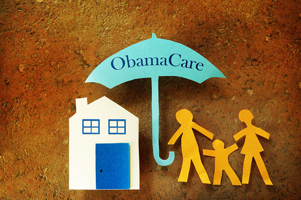 A yellow paper doll next to a green paper umbrella that says ObamaCare next to a white paper house