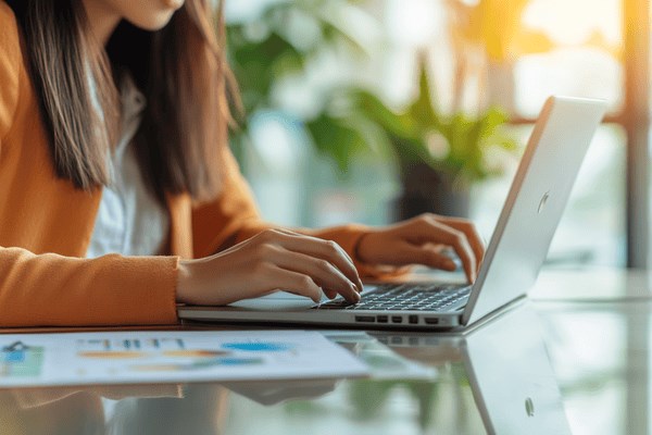 A businesswoman works on a laptop with a document showing graphs on the desk next to her