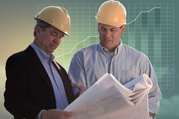 Two men in business clothes and hardhats looking at papers in front of graph