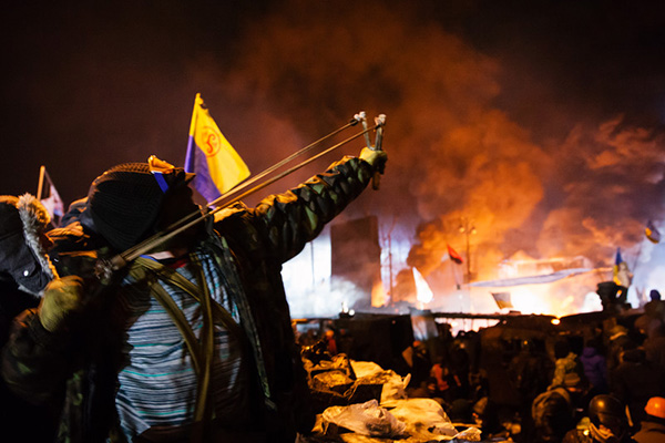 During a riot, a masked rioter is drawing back a slingshot to fire.